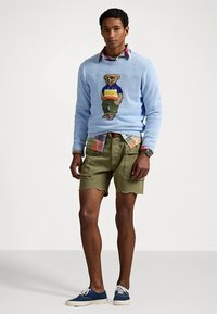 Hellblauer Strickpullover mit Bärengrafik, darüber ein kariertes Hemd getragen. Olivgrüne Shorts und dunkelblaue Segeltuchschuhe vervollständigen den Look.