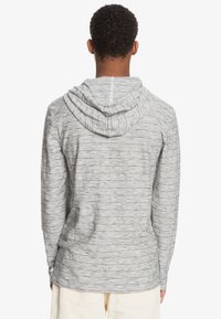 Grå och vit randig långärmad hoodie med dragsnöre i huvan, mjukt material och en något böjd fåll, sedd bakifrån.