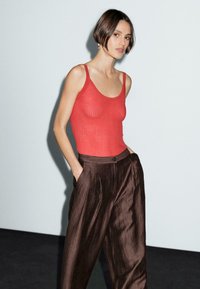 Red ribbed tank top with thin straps paired with wide brown pleated trousers. Smooth texture and form-fitting design. Neutral backdrop.