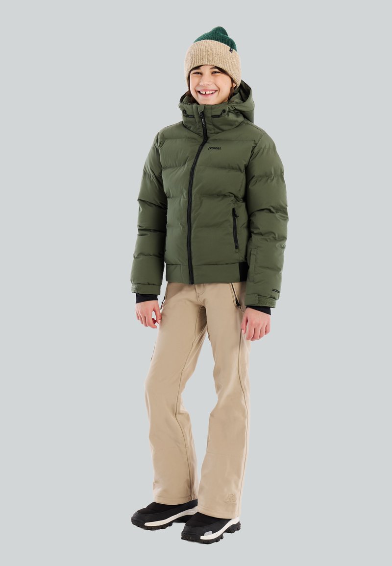 Glunderend kind in een groene pufferjack, beige broek, zwarte sneeuwlaarzen en een beige-groene gebreide muts, staande tegen een effen achtergrond.