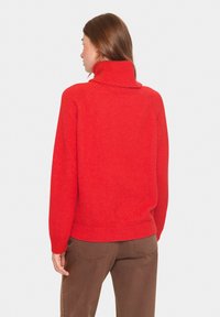 Raudonas vilnonis megztinis su aukštu kaklu, laisvo silueto ir nertomis rankovėmis. Dėvimas su rudos spalvos kelnėmis, parodantis paprastą, tekstūrinį dizainą.