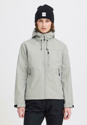 Ženska, oblečena v svetlo sivo jakno z zadrgo in kapuco ter črno kapico, stoji na enostavni svetli podlagi.