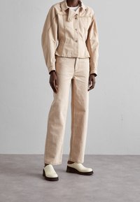Veste en denim beige avec fermeture à bouton et poches avant, associée à un jean droit beige assorti. Sabots blancs avec semelles marron.