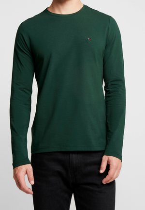 Mężczyzna ubrany w ciemnozieloną koszulkę z długim rękawem i okrągłym dekoltem z małym czerwono-biało-niebieskim logo na lewej piersi, w połączeniu z czarnymi spodniami.