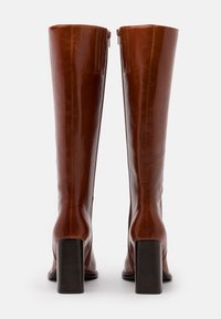 Bottes en cuir marron montantes, avec une finition lisse, des talons carrés et des zips latéraux pour la fermeture. Accentue par des détails de couture minimalistes.