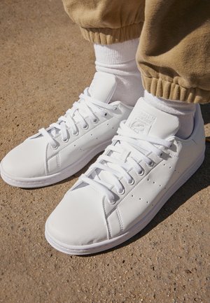 Hvide læder sneakers med snørebånd, båret med hvide sokker og beige bukser, stående på struktureret jord.