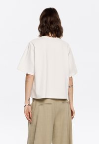 Biały, skrócony t-shirt z krótkimi rękawami, okrągłym dekoltem i miękką teksturą. W zestawie z beżowymi spodniami o szerokich nogawkach z tylnymi kieszeniami.