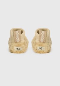Beige UGG slippers med myk, teksturert pels på overdelen, strikket hælflik, og et logopatch bak. Hælen har et dekorativt sikksakkmønster.