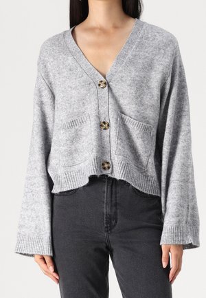 Femme portant un cardigan en maille gris clair à trois boutons sur le devant et deux poches, associé à un pantalon taille haute gris foncé.