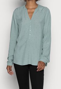 Blouse vert clair à manches longues avec un col en V, dotée d'une patte de boutonnage et de petits pois noirs. Tissu doux avec une coupe décontractée.
