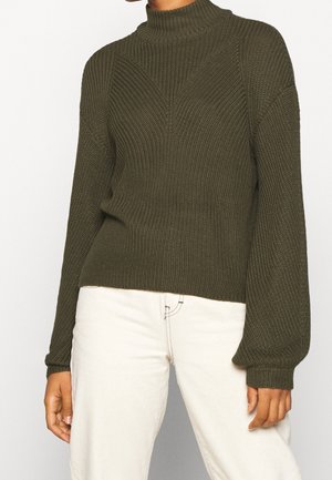 Personne portant un pull à col roulé côtelé vert olive avec des manches ballon et un pantalon taille haute ivoire.