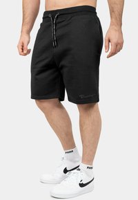 Shorts neri in cotone con una cintura elastica, chiusura con stringa e un piccolo logo sull'orlo. Abbinati a sneakers bianche e calzini alla caviglia.