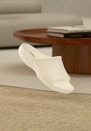 Hvite slip-on tresko sandaler med ventilasjonshull, tykk såle, plassert nær et trebord med bøker i en stue.