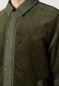 Veste en velours côtelé vert olive avec boutons-pression, tissu texturé, col et texte brodé discret sur la poitrine gauche.