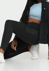 Donna con leggings neri Adidas, reggiseno sportivo blu chiaro, giacca nera e sneakers bianche, inginocchiata con una mano sul pavimento.