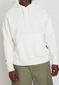 Bílá mikina s kapucí, přední kapsou a šňůrkou na stažení. Rukávy typu raglan a štítek s logem na boku. Měkká textura.