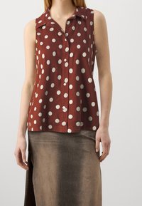 Blusa marrón sin mangas con lunares blancos, diseño con cuello y cierre frontal de botones. Hecha de una tela ligera con un ajuste relajado.