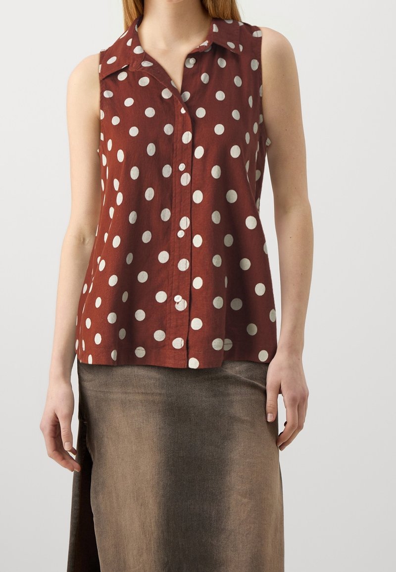 Blusa marrón sin mangas con lunares blancos, diseño con cuello y cierre frontal de botones. Hecha de una tela ligera con un ajuste relajado.