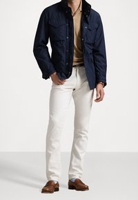 Marinblå lättviktsjacka med dragkedja och tryckknappar, två framfickor, beige tröja, vita tajta byxor, bruna loafers med ljusblå strumpor.