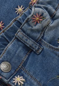 Denimtyg med färgglad blommönstring i rosa, gult och grönt. Inkluderar en silverfärgad metallknapp och kontrasterande sömdetaljer.