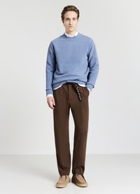 Junger Mann in einem hellblauen Pullover über einem weißen Hemd, braunen Hosen mit einem geflochtenen Gürtel und beigen Wildlederschuhen, der vor einem einfarbigen Hintergrund steht.