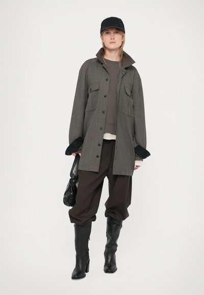 Mujer vestida con una chaqueta grande gris, pantalones amplios oscuros, botas negras, gorra negra y sosteniendo un bolso negro contra un fondo blanco.