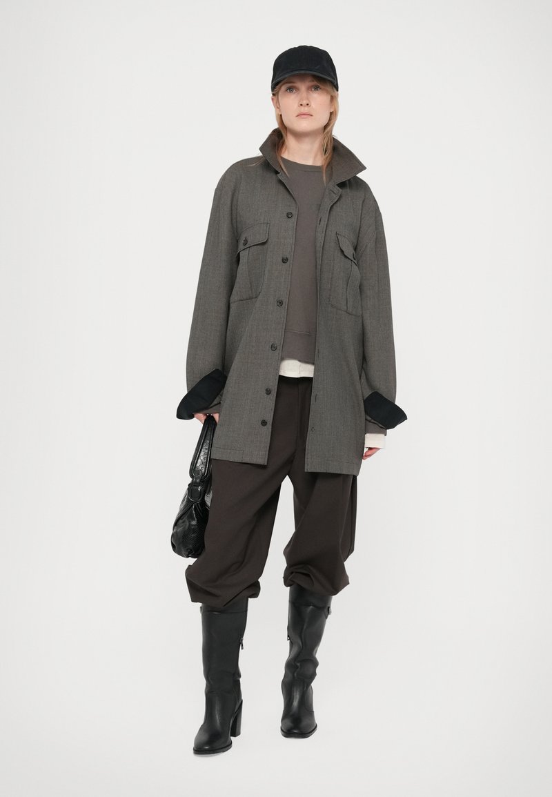 Femme portant une veste grise oversized, un pantalon baggy noir, des bottes noires, une casquette noire, et tenant un sac à main noir sur fond blanc.