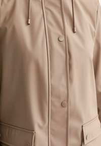 Impermeabile beige con bottoni a pressione, due tasche frontali con patta e cappuccio con coulisse visibile nella parte superiore del petto e in vita.