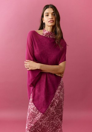 Femeie cu părul lung și castaniu, purtând un șal mov peste o rochie roz cu imprimeu floral, stând cu brațele încrucișate pe un fundal roz.