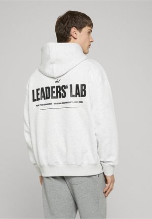 Junger Mann im Seitenprofil, trägt einen hellgrauen Hoodie mit schwarzem "LEADERS' LAB" Schriftzug auf dem Rücken und passende graue Jogginghose vor einfarbigem Hintergrund.