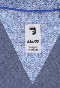 Blauwe en grijze stoffen met een patroon en een driehoekige vorm. Een wit label met de tekst "J&JOY" boven "Ontworpen in België," met een logo.