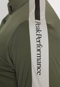 Polo vert olive avec col, bande latérale noire et blanche portant le texte "Peak Performance", et tissu lisse et texturé.