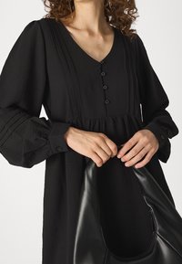 Robe noire à manches longues avec un décolleté en V et une fermeture à boutons ; tissu texturé avec une taille froncée, associée à un sac en cuir noir au design courbé.