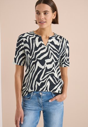 Femme portant un haut à manches courtes rayé zèbre noir et blanc et un jean bleu clair, une main dans la poche, sur fond beige.