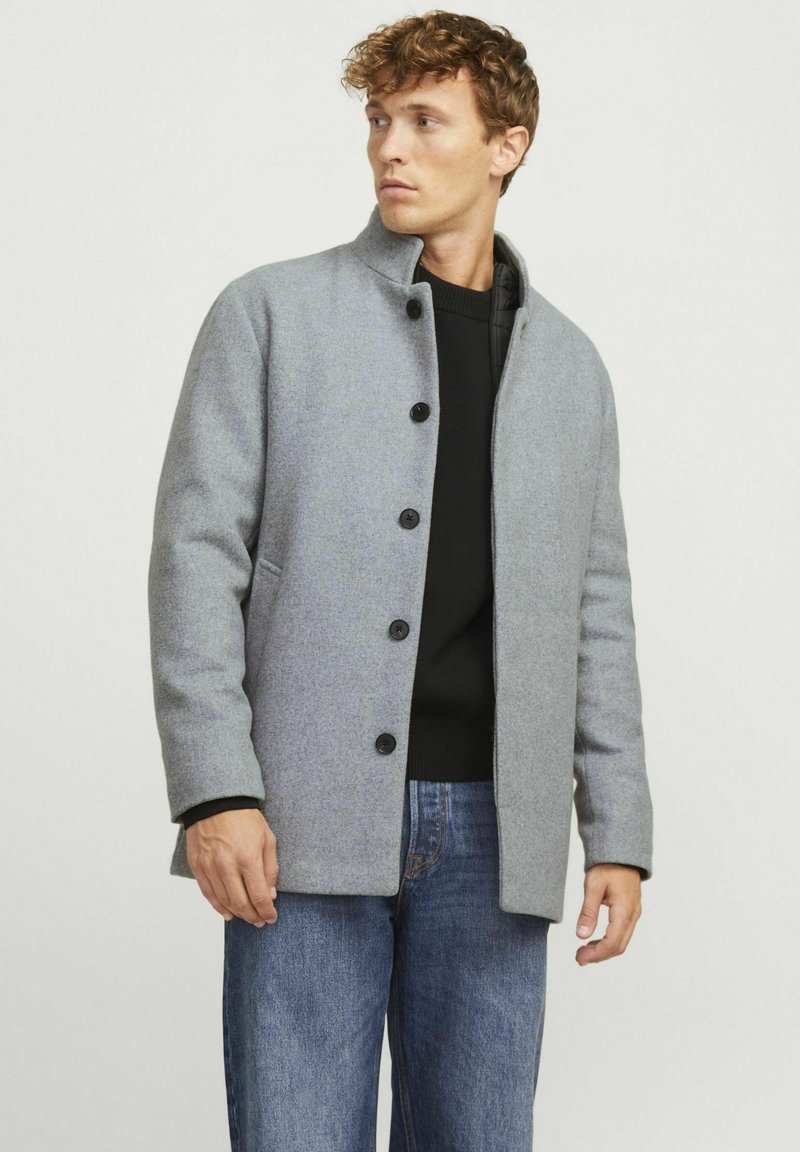 Abrigo de lana gris con un cuello alto, botones en la parte delantera y bolsillos laterales, usado sobre un suéter negro y combinado con unos vaqueros azules. Textura suave.