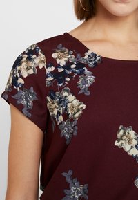 Burgundská blůza s krátkým rukávem, zdobená květinovými vzory v krémové, námořnické a šedé barvě. Měkká tkanina s hladkým povrchem a kulatým výstřihem.