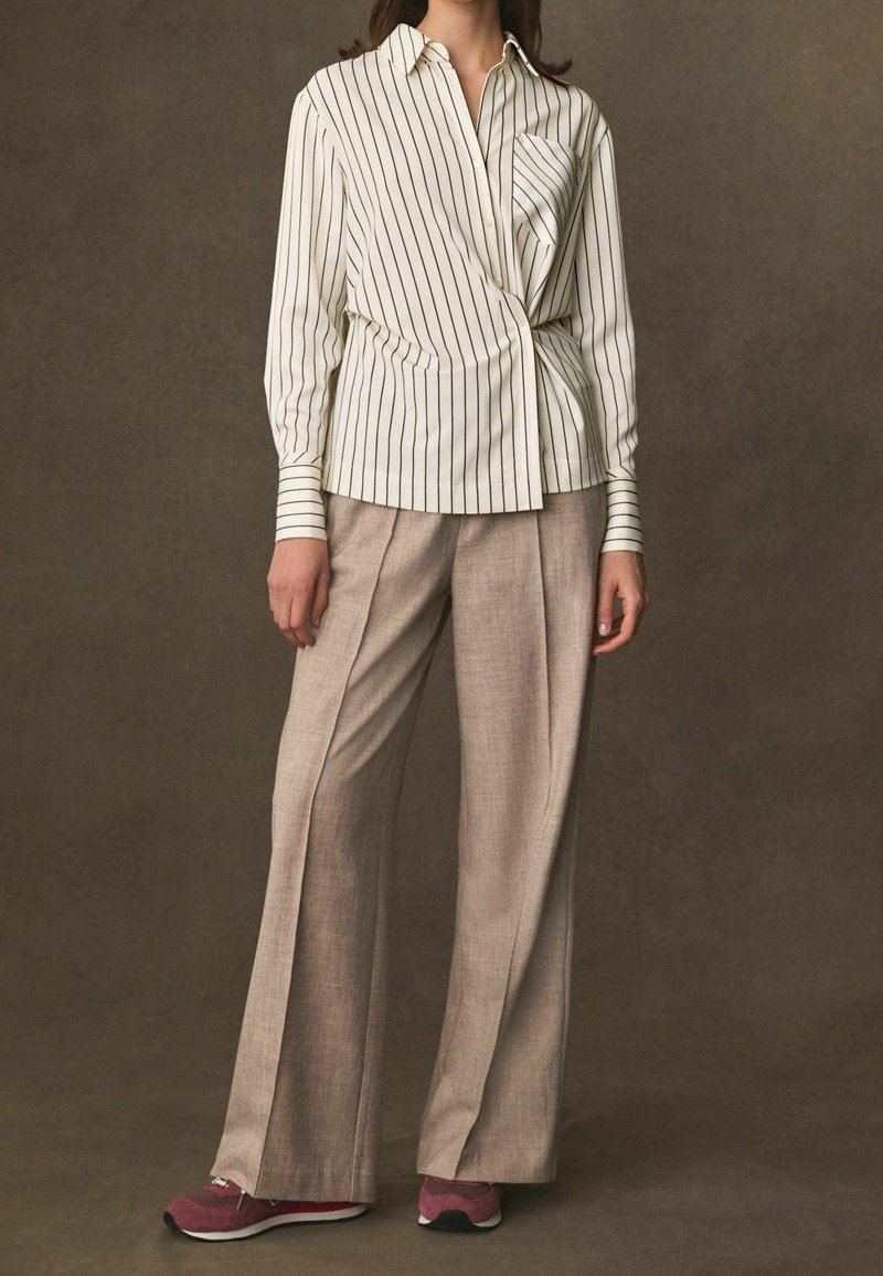 Weiß gestreifte Bluse mit Knotendetail, beigefarbene weite Hose und kastanienrote Sportschuhe. Glattes Material, entspannter Schnitt und geknöpfte Manschetten.