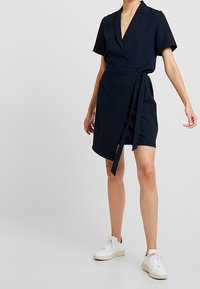Mujer vestida con un vestido cruzado de manga corta azul marino con un cinturón y zapatillas blancas, de pie contra un fondo claro plano.