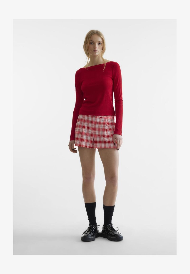 Rotes gestreiftes Langarmoberteil, kombiniert mit einem karierten Minirock in Weiß-, Rot- und Hellrosa-Tönen. Schwarze kniehohe Socken und klobige Schuhe.