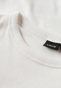 Valkoinen ribbineulekangas, jossa on pyöreä kaula-aukko, ja kauluksessa musta etiketti, jossa lukee "LMTD". Pehmeä rakenne hienoilla viivoilla.