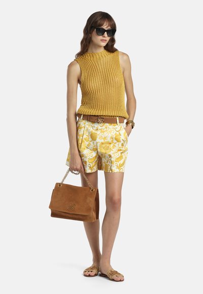 Femme portant des lunettes de soleil noires, un haut en tricot sans manches moutarde, un short blanc à motifs jaunes, une ceinture marron, des sandales et tenant un sac à main en daim marron.