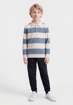 Garçon blond debout les mains détendues, portant une chemise à manches longues rayée bleu et beige, un pantalon noir et des baskets blanches sur fond blanc.
