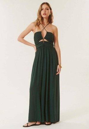 Femme portant une robe longue vert foncé à col halter avec des découpes et des sandales noires, debout avec une main sur la hanche.