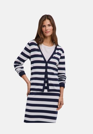 Donna che indossa un cardigan a righe orizzontali blu navy e bianco e una gonna abbinata, posa con una mano sul fianco davanti a uno sfondo semplice.