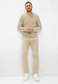 Beige Strickpullover mit strukturierten Details, kombiniert mit hellbeigen Cordhosen und weißen Sneakern, vor einem schlichten weißen Hintergrund stehend.