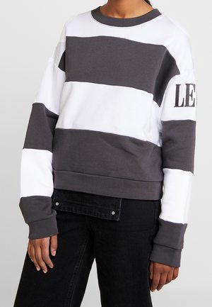Personne portant un sweat-shirt à manches longues et col rond avec de larges rayures horizontales blanches et grises et un texte partiel "LE" sur la manche gauche.