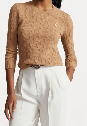 Kvinde iført en kamelfarvet kabelstrikket sweater kombineret med højtaljede hvide bukser og et brunt bælte med en dekorativ spænde.