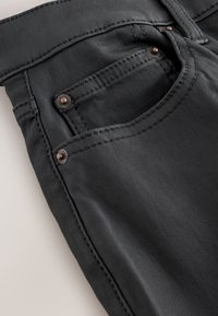 Crne traperice s glatkom teksturom, s prednjim džepom, bakrenim metalnim detaljima i kontrastnim šavovima duž šavova.