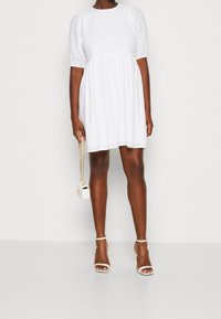 Vestido blanco de manga abullonada con un patrón texturizado de lunares, cintura ceñida y un corte relajado, combinado con sandalias de tacón blancas y un bolso pequeño.