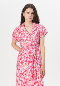 Blomstertrykt kjole i nuancer af pink og rød. Korte ærmer, V-halsudskæring, og en båndsløjfe i taljen. Glat stof med en blød tekstur.